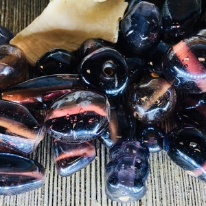 May include: A collection of dark purple and brown glass beads with a shiny, iridescent finish. The beads are various shapes and sizes, including round, oval, and rectangular.