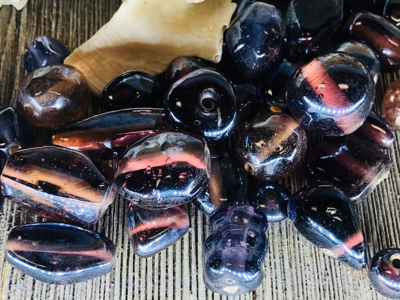 May include: A collection of dark purple and brown glass beads with a shiny, iridescent finish. The beads are various shapes and sizes, including round, oval, and rectangular.