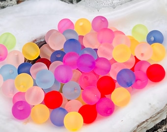 May include: A collection of colorful translucent round beads in various shades of pink, blue, yellow, red, and green. The beads are arranged in a pile on a white surface.