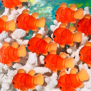 May include: A group of orange and yellow clownfish figurines are arranged on a white coral reef background. The figurines are made of a translucent plastic material and have a realistic appearance.