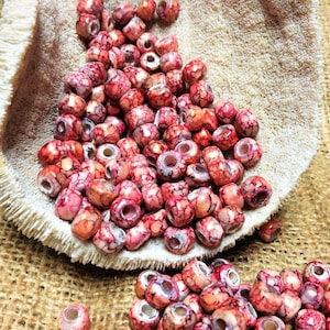 May include: A close-up of a pile of red and white speckled beads. The beads are round and have a hole in the center. They are in a white bowl and some are spilling out onto a brown burlap surface.