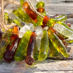 May include: A collection of glass beads in shades of green, brown, and clear. The beads are shaped like long, thin teardrops and are strung together on a twine thread.