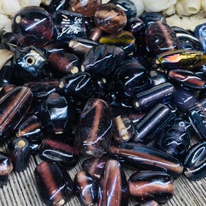 May include: A collection of dark purple and black glass beads with various shapes and sizes. Some beads have a shiny, iridescent finish, while others have a matte finish. The beads are perfect for jewelry making or crafting.
