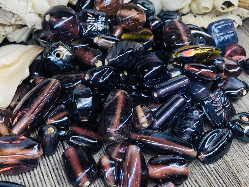 May include: A collection of dark purple and black glass beads with various shapes and sizes. Some beads have a shiny, iridescent finish, while others have a matte finish. The beads are perfect for jewelry making or crafting.