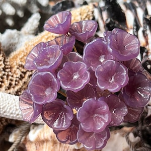 以下が含まれることがあります： 繊細な虹色の仕上げが施された、小さな紫色のガラスの花の集まり。花はブーケのような形にアレンジされており、手作りのようです。