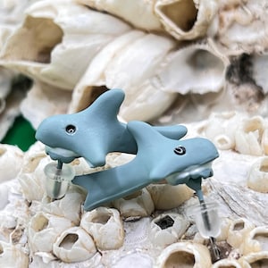 May include: A pair of light blue shark-shaped stud earrings. The earrings have a small black eye and a white mouth. The earrings are set against a background of white and beige barnacles.