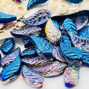 May include: A collection of iridescent blue leaf-shaped beads. The beads have a glossy finish and intricate vein detailing. Some beads have a silver or gold sheen, creating a shimmering effect. The beads are scattered on a white surface.