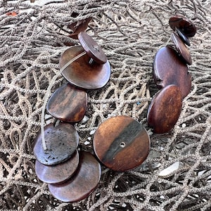 May include: A necklace made of dark brown wooden beads, some round and some oval, strung on a clear cord. The beads are arranged in a staggered pattern, with the round beads at the top and the oval beads at the bottom. The necklace is lying on a beige net-like surface.