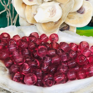 Puede incluir: Una colección de cuentas de vidrio translúcidas de color rubí, exhibidas en una gran concha blanca. Las cuentas son redondas y cilíndricas, con agujeros visibles para ensartar. El fondo incluye una red verde y blanca.