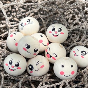 Peut inclure: Un ensemble de perles en bois avec différents visages de dessins animés mignons peints dessus. Les perles sont blanches avec des yeux noirs et des joues roses. Certains des visages comprennent un chat, un cœur, un sourire et un clin d'œil.