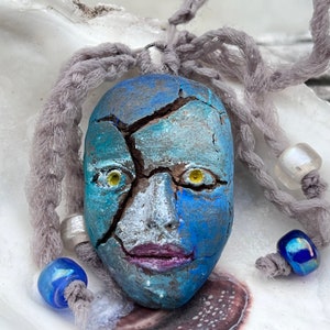 Puede incluir: Un colgante de cerámica azul y blanco con una cara agrietada y ojos amarillos. El colgante está unido a un cordón gris con cuentas azules y blancas.