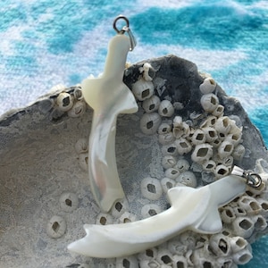 Peut inclure: Deux pendentifs en nacre blanche en forme de poisson, avec des anneaux de saut en argent. Les pendentifs reposent sur une surface texturée blanche et grise.