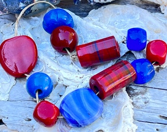 Peut inclure: Un collier composé de perles rouges et bleues de différentes formes. Certaines perles sont rayées, d'autres rondes ou carrées. Les perles sont enfilées sur un cordon naturel. Le collier est présenté sur une surface blanche texturée.