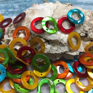 May include: Assortment of colorful iridescent shell rings, with a variety of colors including yellow, green, blue, and red. The rings are arranged on a white seashell background.