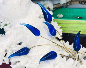 Könnte beinhalten: Eine Gruppe von blauen Glas-Tropfenornamenten auf dünnen goldenen Drähten, angeordnet auf einer weißen korallenartigen Oberfläche.