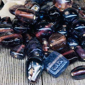 May include: A collection of dark purple glass beads with various shapes and sizes. The beads have a translucent quality and some have a slight iridescent sheen.