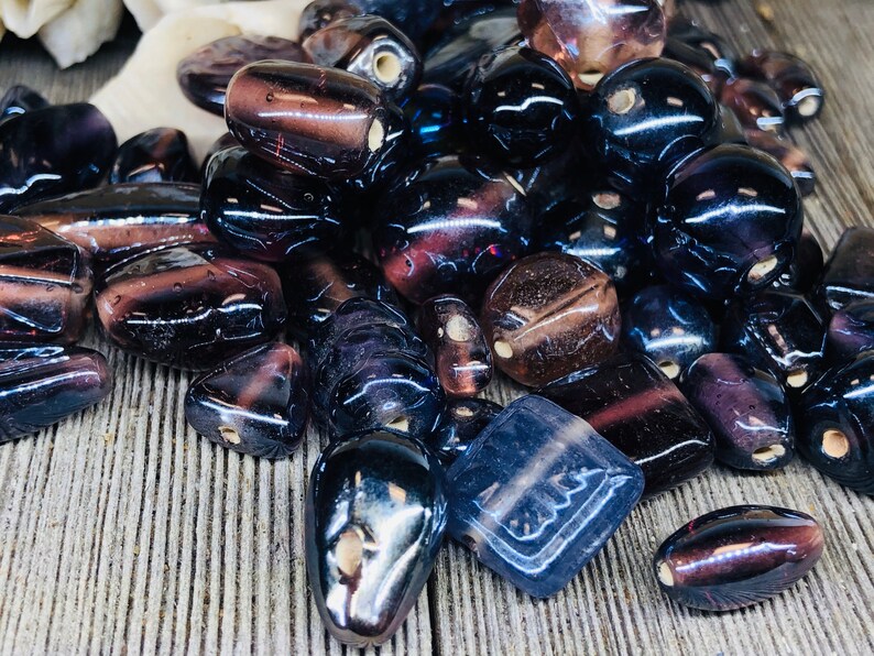 May include: A collection of dark purple glass beads with various shapes and sizes. The beads have a translucent quality and some have a slight iridescent sheen.