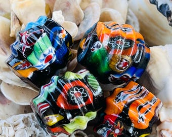 Peut inclure: Quatre perles de verre colorées et à motifs avec une base noire. Les perles présentent des motifs complexes dans les tons de bleu, vert, orange, rouge et blanc. Les perles sont exposées sur un lit de coquillages.
