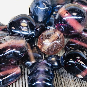 May include: A collection of purple and clear glass beads with various shapes and sizes. The beads have a glossy finish and are arranged on a wooden surface.