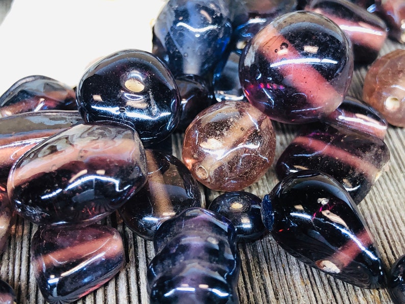 May include: A collection of purple and clear glass beads with various shapes and sizes. The beads have a glossy finish and are arranged on a wooden surface.