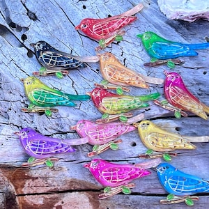 May include: A collection of 12 colourful bird-shaped glass ornaments with gold accents. The ornaments are arranged on a wooden surface and feature a variety of colours, including red, blue, green, yellow, purple, and pink.