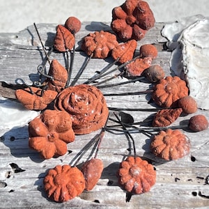 Puede incluir: Una colección de formas de flores y hojas de terracota en tallos de alambre. Las formas son de varios tonos de rojo y marrón, y están dispuestas en un patrón circular sobre una superficie de madera desgastada.