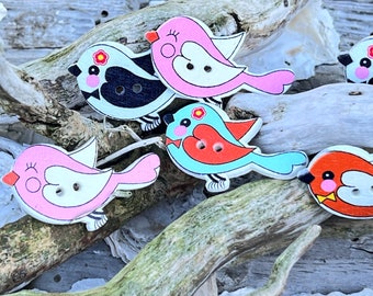 May include: Six wooden bird buttons with painted details. The birds are in various colors, including pink, blue, red, and black. Each bird has a white face with black eyes and a small flower on its chest.