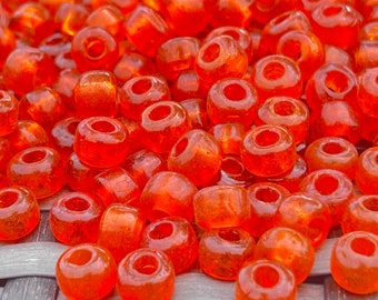May include: A close-up of a pile of small, orange glass beads. The beads are round and have a hole in the center. They are arranged in a random pattern.