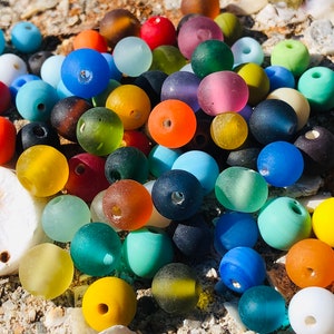 Może przedstawiać: Kolekcja kolorowych szklanych koralików, niektóre z matowym wykończeniem, w różnych odcieniach niebieskiego, zielonego, żółtego, pomarańczowego, czerwonego i fioletowego. Korale są rozrzucone na piaszczystej powierzchni.