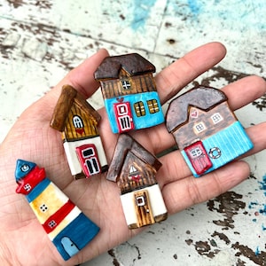 May include: Five small, hand-painted ceramic house and lighthouse figurines. The collection includes four houses with brown roofs, white or blue walls, and red or brown doors, plus one tall lighthouse with blue, white, and red horizontal stripes.