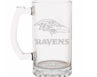 May include: A clear glass beer mug with a handle. The mug features the Baltimore Ravens logo and the word "RAVENS" in gray. The mug has a rectangular shape and a decorative base.