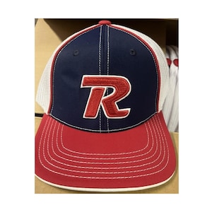 May include: A baseball cap with a navy blue crown and a red bill. The front of the cap features a large, embroidered red "R" with a white outline. The back of the cap is white mesh.