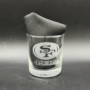 May include: A clear glass tumbler with the San Francisco 49ers logo etched on the front. The logo is a silver oval with "SF" inside and "49ERS" below. A black cloth is inside the glass.