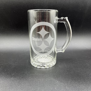 K&ouml;nnte beinhalten: Klarer Bierkrug aus Glas mit Henkel. Der Krug zeigt das Logo der Pittsburgh Steelers, ein Design mit drei Diamanten in einem Oval, mit dem Wort "Steelers" darunter. Der Krug hat einen dekorativen Boden.