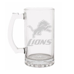 May include: Clear glass beer mug with a handle. The mug features the Detroit Lions logo and the word "Lions" in gray. The mug has a rectangular shape and a decorative base.