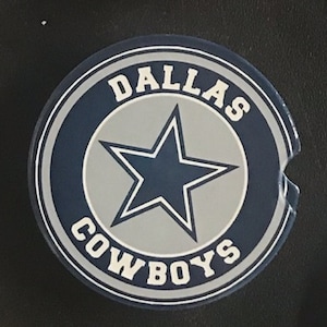 May include: A round car coaster featuring the Dallas Cowboys logo. The coaster has a navy blue border with the words "DALLAS COWBOYS" in white. The center has a gray background with a navy blue star.