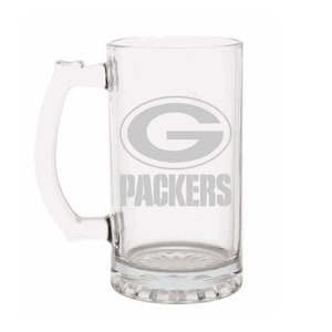 May include: Clear glass beer mug with a handle. The mug features the Green Bay Packers logo and the word "PACKERS" in gray. The mug has a decorative base.