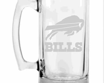 May include: Clear glass beer mug with a handle. The mug features the Buffalo Bills logo and the word "BILLS" in gray. The mug is rectangular with a thick base and a curved handle.