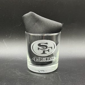 May include: A clear glass tumbler featuring the San Francisco 49ers logo etched on the front. The logo is a stylized "SF" inside an oval. The words "49ERS" are etched below the logo. A black cloth napkin is inside the glass.