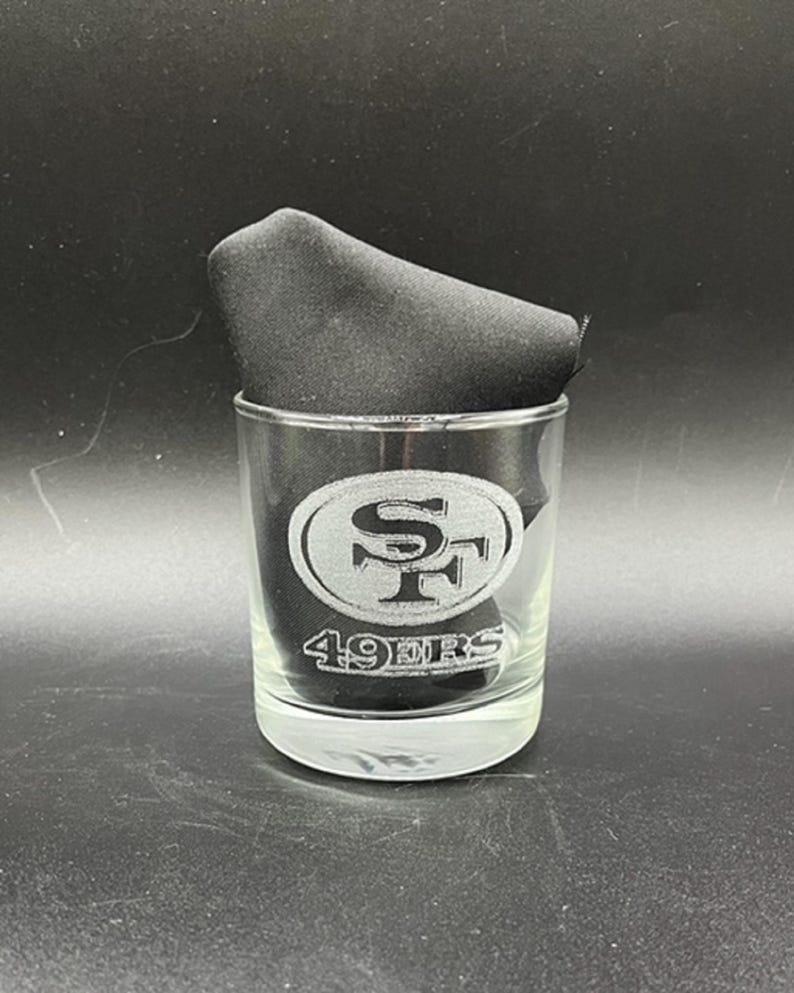 May include: A clear glass tumbler featuring the San Francisco 49ers logo etched on the front. The logo is a stylized "SF" inside an oval. The words "49ERS" are etched below the logo. A black cloth napkin is inside the glass.
