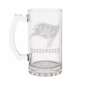 May include: Clear glass beer mug with a handle. The mug features the Tampa Bay Buccaneers logo, a skull and crossbones with a football, and the word "BUCCANEERS" in gray.