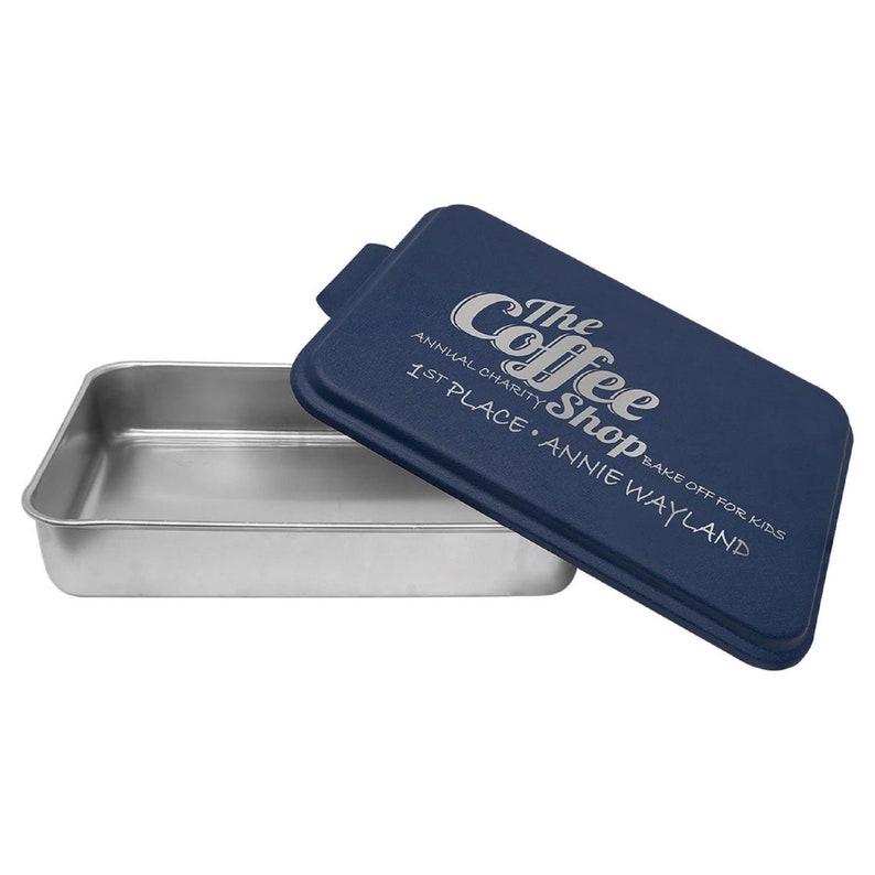 May include: A blue baking pan with a lid. The lid has a white design that says "The Coffee Shop" with the text "Annual Charity Bake Off for Kids 1st Place Annie Wayland".