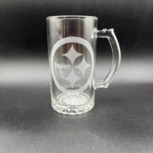 May include: Clear glass beer mug with a handle. The mug features the Pittsburgh Steelers logo, a three-diamond design inside an oval, with the word "Steelers" below. The mug has a decorative base and is suitable for beverages.