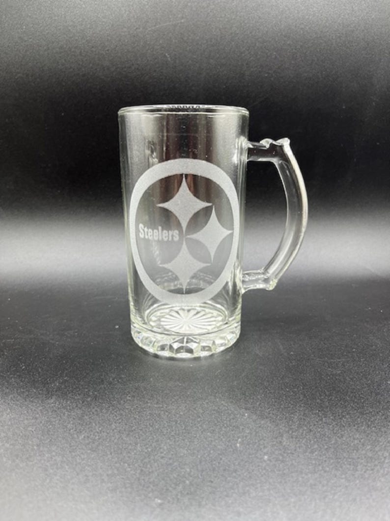 May include: Clear glass beer mug with a handle. The mug features the Pittsburgh Steelers logo, a three-diamond design inside an oval, with the word "Steelers" below. The mug has a decorative base and is suitable for beverages.