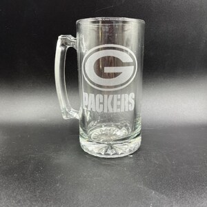 May include: Clear glass beer stein with a handle. The stein features the Green Bay Packers logo and the word "PACKERS" etched on the front. The base of the stein has a decorative, textured design.