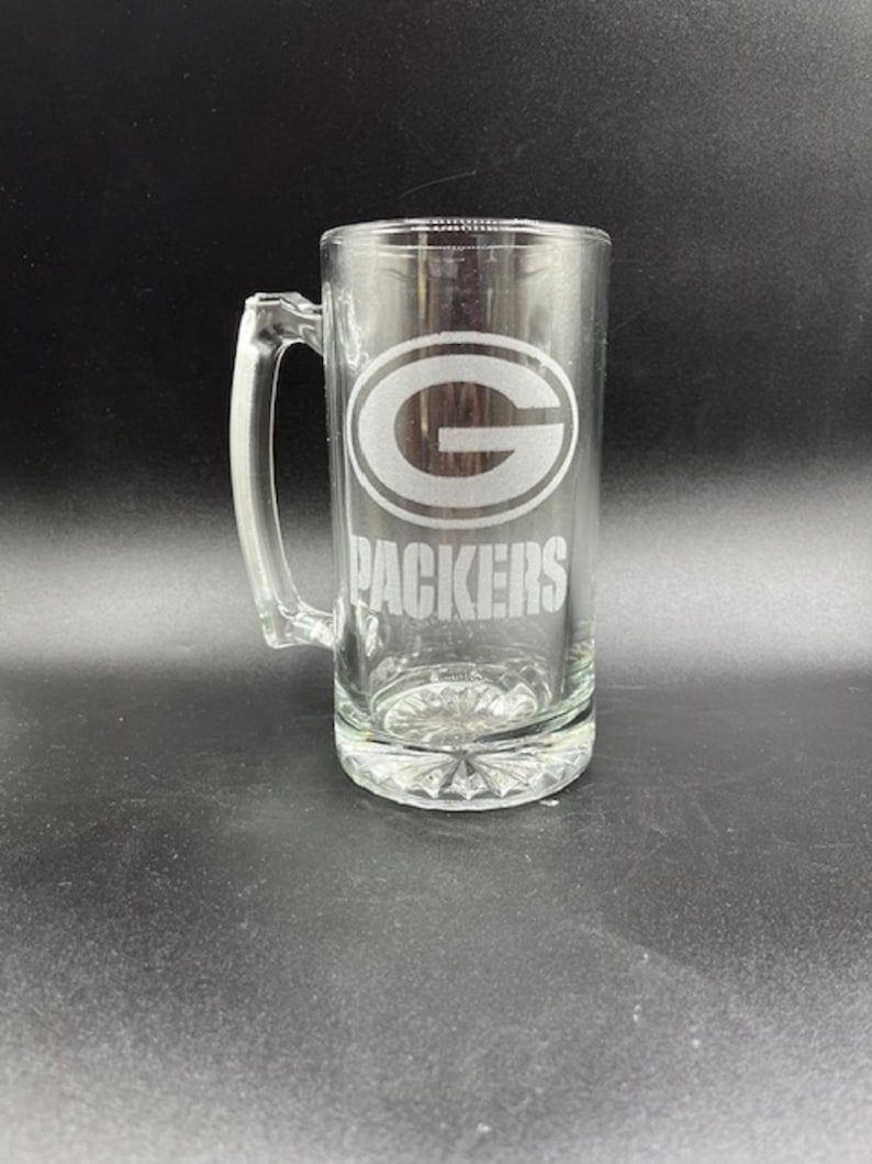 May include: Clear glass beer stein with a handle. The stein features the Green Bay Packers logo and the word "PACKERS" etched on the front. The base of the stein has a decorative, textured design.