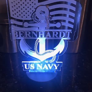 May include: A white acrylic night light with a US Navy anchor design. The anchor is surrounded by a rope design and a US flag. The name "BERNHARDT" is written above the anchor and "US NAVY" is written below the anchor.