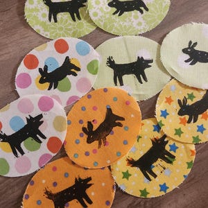 May include: A collection of circular fabric patches with black dog illustrations. The patches feature various patterns, including polka dots, floral designs, and stars, in colors like green, orange, and yellow. The dog illustrations are hand-drawn.