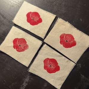 May include: Four square, beige fabric coasters with a red poppy flower design. The poppies are centered on each coaster, with a black center. The coasters have a frayed edge.