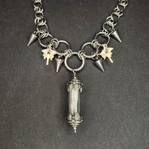 May include: A silver-toned necklace with a chain link design. The necklace features cone-shaped pendants, small bone charms, and a cylindrical pendant with a decorative cap. The background is a dark gray.